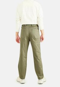 Olijfgroene broek van stevig materiaal, met een recht model, achterzakken en een tailleband met riemlussen. Schoenen in een lichte kleur.