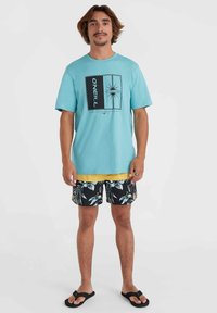 Camiseta turquesa con un diseño gráfico, combinada con pantalones cortos amarillos y bañadores negros con un patrón de hojas tropicales. Chanclas negras.
