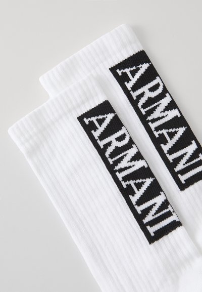 Calcetines blancos de canalé con un logo rectangular negro que dice "ARMANI" en blanco, situado de manera prominente en el lateral. Textura suave.