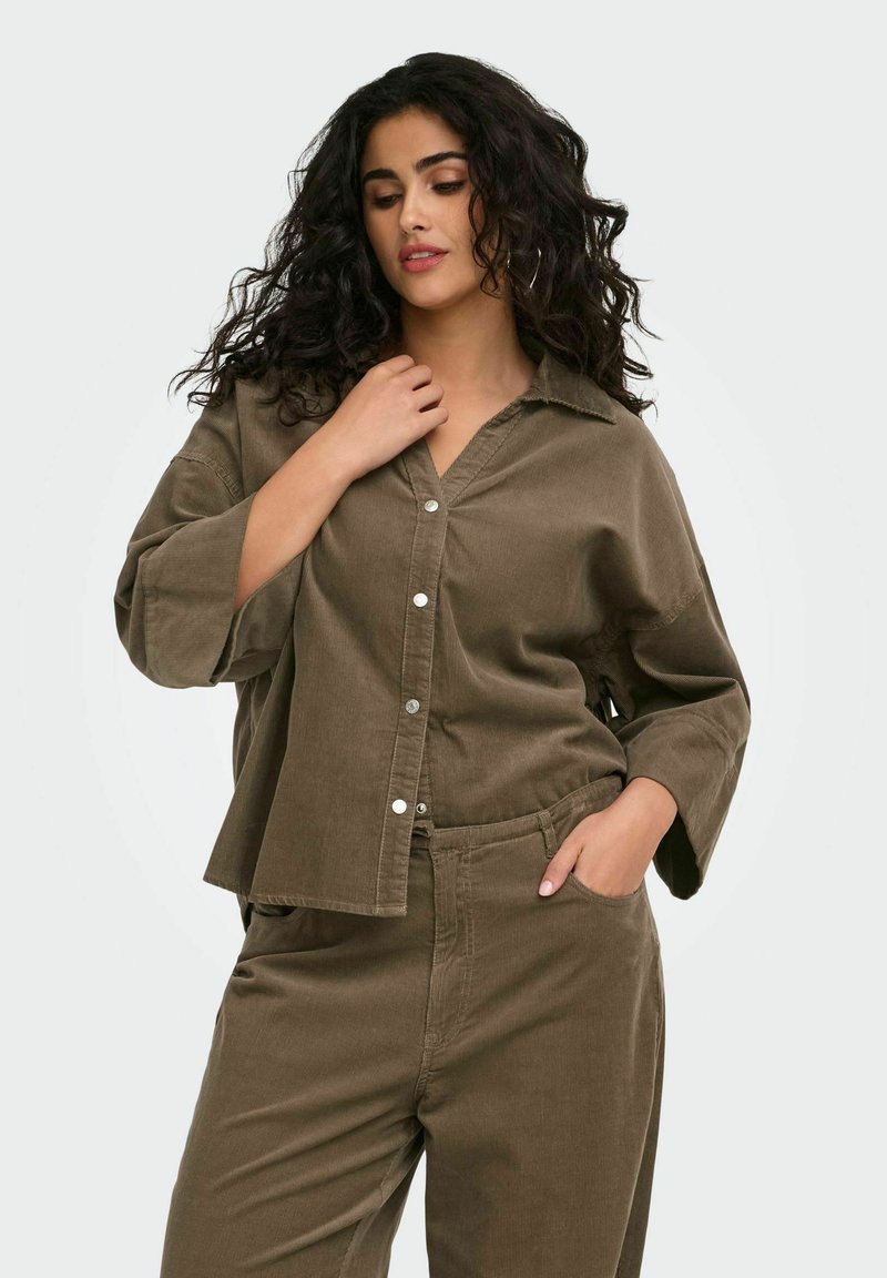 Femme aux cheveux bouclés et foncés portant une chemise marron ample à boutons et un pantalon assorti, posant avec une main près du col et une autre dans la poche.