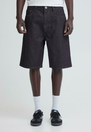 Man draagt zwarte spijkershorts, witte sokken en zwarte sneakers, staat tegen een lichte effen achtergrond.