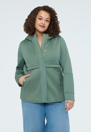 Nainen kiharatukka päällään vihreä, kohokuvioitu vetoketjullinen ja neppareilla varustettu takki, yhdistetty vaaleansinisiin farkkuihin, yksiväristä taustaa vasten.