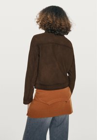 Bruine suède cropped jas met schoudernaden, gedragen over een oranje laagjes kledingstuk. Gerenoveerde afwerking en comfortabele pasvorm.