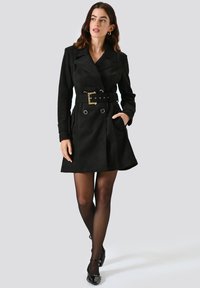 Cappotto in suede nero con cintura, colletto a revers, design doppiopetto e dettagli in occhio di chiave argentati. Lungo fino al ginocchio con tasche laterali.
