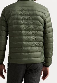 Mężczyzna w oliwowej, pikowanej kurtce puchowej oraz ciemnoszarych spodniach, pokazany od tyłu na tle gładkiej, białej ściany.