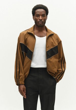Homme portant une veste zippée marron et noire, un débardeur blanc et un pantalon noir, debout et regardant devant sur un fond uni.