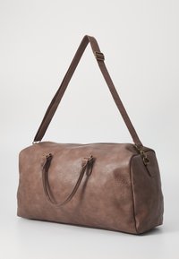 Brun lær duffel bag med teksturert overflate, doble håndtak, justerbar stropp og messingdetaljer. Rektangulær form.