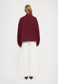 Bordeaux wollen trui met een hoge kraag en een oversized pasvorm, met verlaagde schouders, gecombineerd met witte wijde broek.