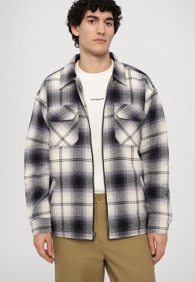 Plaid jacket in white, purple, and black, featuring two front pockets, a zipper, and a warm, textured fabric. Worn over a white shirt.