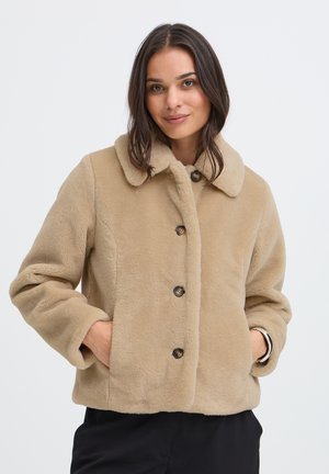 Femme portant une veste beige pelucheuse boutonnée avec un col, les mains dans les poches et un pantalon noir, debout devant un fond clair uni.
