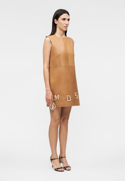 Γυναίκα στέκεται φορώντας αμάνικο δερμάτινο φόρεμα ταμπά με λευκά γράμματα από σκοινί κοντά στο τελείωμα, συνδυασμένο με μαύρα πέδιλα με λουριά, σε απλό φόντο.