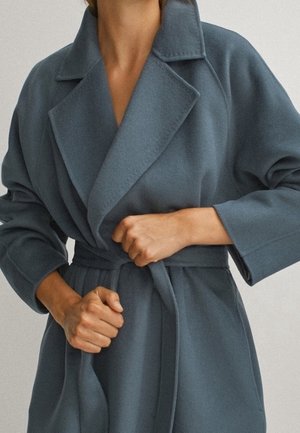 Personne attachant la ceinture d'un long manteau en laine bleu-gris avec de larges revers et de longues manches.