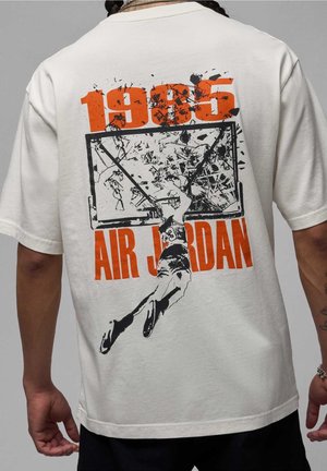 Personne portant une chemise blanche avec un graphique d'un joueur de basket dunkant à travers un panneau arrière brisé, avec les textes "1985" et "Air Jordan" en orange.