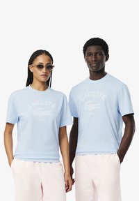 Camisetas de algodón azul claro con mangas cortas, que presentan un logo de cocodrilo blanco y el texto "PARÍS". Combinadas con pantalones de chándal rosa pálido.