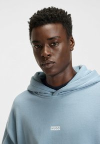 Lichtblauwe hoodie ontworpen met een textuurstof, voorzien van een logo op de voorkant en een dubbele capuchon. De ronde hals heeft meerdere lagen en is ontspannen.
