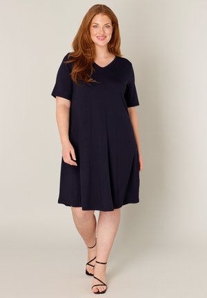 Vrouw met lang rood haar, gekleed in een donkerblauwe jurk tot op de knie met korte mouwen en zwarte sandalen met bandjes, staand tegen een effen achtergrond.