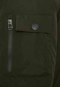 Prenda exterior verde con un bolsillo con cremallera y un bolsillo con solapa con un botón negro. El material tiene una textura lisa y mate.