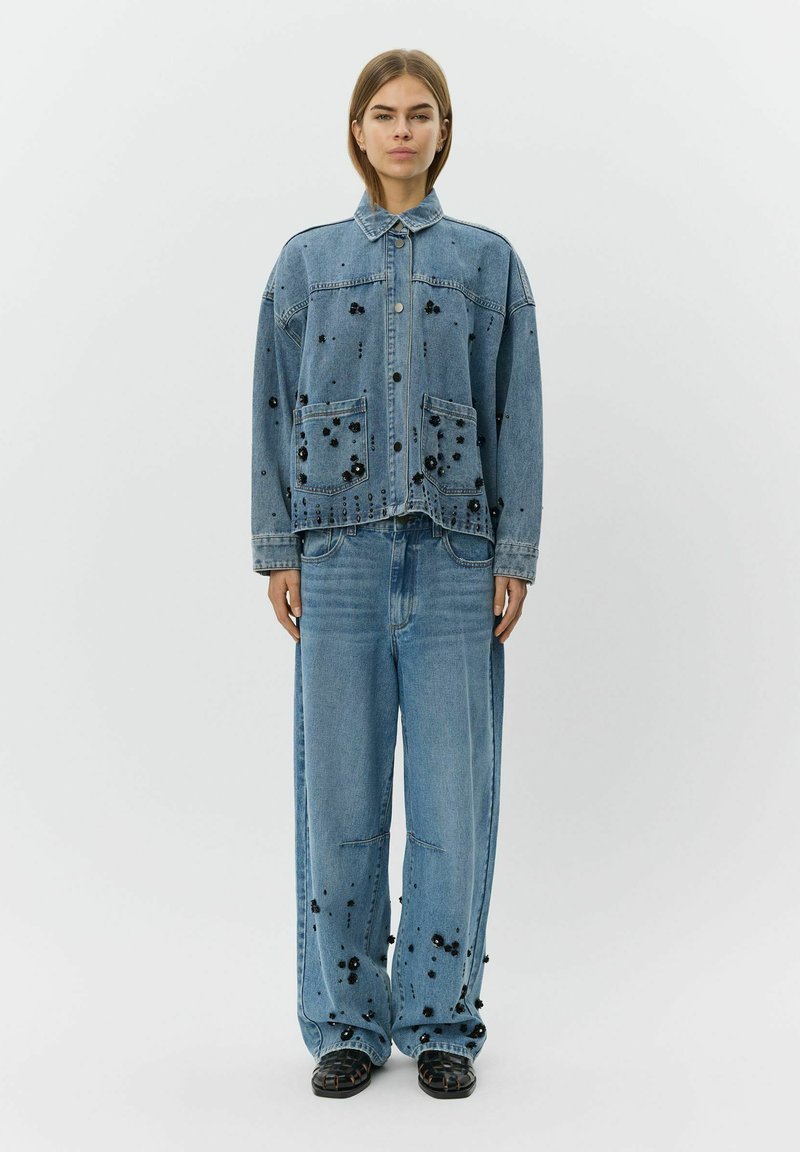 Lyseblå denimjakke og matchende bukser med sorte pyntedetaljer og oversized pasform. Jakken har to forlommer og knaplukning.