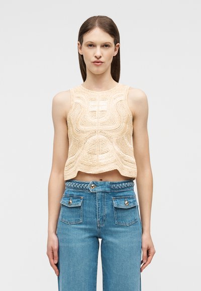 Femme portant un crop top beige en crochet sans manches et un jean bleu taille haute avec poches avant à rabat et ceinture tressée.