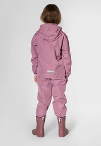 Różowa wodoodporna kurtka z kapturem i dopasowane spodnie, pokazane od tyłu. Posiadają elastyczne mankiety oraz brązowe gumowe kalosze. Prosty design.