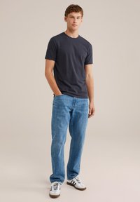 Tânăr stând îmbrăcat cu un tricou bleu închis, blugi deschiși la culoare și adidași albi cu dungi negre, pe un fond simplu.