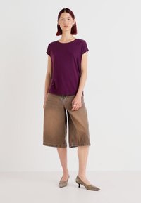 Lilla kortermet t-skjorte kombinert med løse brune culottes og dempede hæler, satt mot en ensfarget hvit bakgrunn.