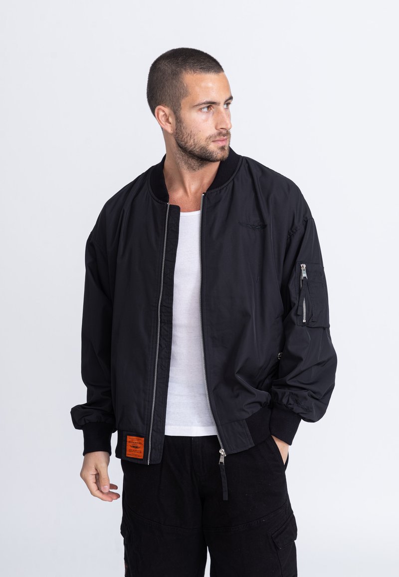 Schwarze Bomberjacke mit Reißverschluss, gerippten Bündchen und einem orangefarbenen Etikett. Verfügt über Seitentaschen und eine glatte Stoffoberfläche.
