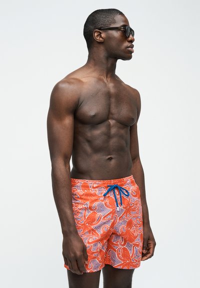 Μυώδης άνδρας με μαύρα γυαλιά ηλίου φορώντας πορτοκαλί μαγιό με σχέδια από χταπόδι, στέκεται μπροστά σε απλό ανοιχτό φόντο.