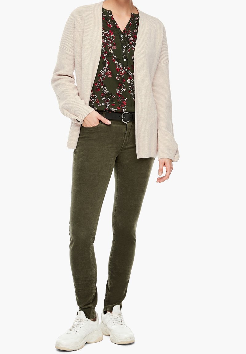 Femme portant un gilet beige, un chemisier à fleurs vert foncé, un pantalon vert olive, une ceinture noire et des baskets blanches, debout avec une main dans la poche.
