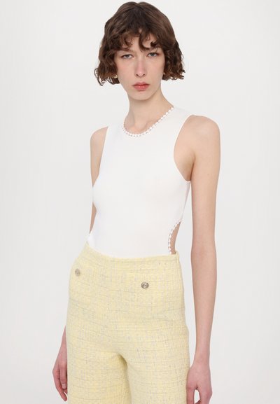 Valkoinen ilmanhiha bodysuit, jossa on aallotettu kaula-aukko, yhdistettynä korkea vyötäröinen keltainen tweed-housu, jossa on kaksi koristepainiketta.