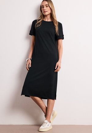 Kobieta stoi przy ścianie, ubrana w czarną sukienkę midi z podwiniętymi krótkimi rękawami i białe trampki.