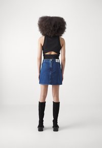 Kvinna med lockigt hår står vänd bort, klädd i en svart ärmlös topp med urklipp, jeanstygkjol och svarta knähöga stövlar mot vit bakgrund.