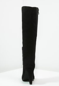 Botte haute en suède noir avec un talon effilé, présentant une fermeture éclair sur le côté et des accents métalliques décoratifs le long de la couture arrière.