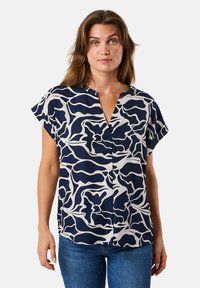 Navy- und cremefarbenes kurzärmliges Bluse mit abstraktem Wellenmuster, V-Ausschnitt und lockerer Passform, kombiniert mit blauen Jeans. Weicher Stoff.
