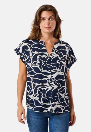 Blusa corta a maniche corte navy e crema con motivo astratto a onde, scollatura a V e vestibilità ampia, abbinata a jeans blu. Tessuto dalla consistenza morbida.