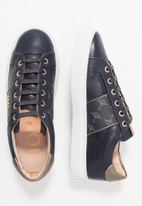 Baskets en cuir bleu marine avec semelle en caoutchouc blanche. Elles présentent des œillets en or rose, un motif décoratif sur les côtés et des accents couleur tan sur la languette et le talon.