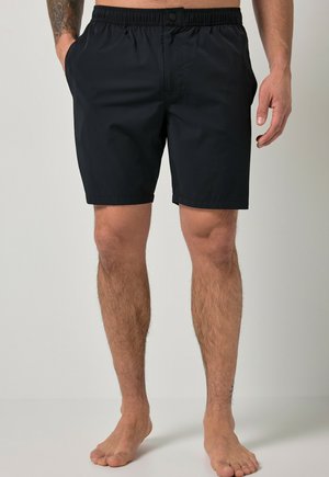 Mand iført sorte elastiske shorts står barfodet mod en ensfarvet baggrund, med delvist synlig tatoveret venstre arm.