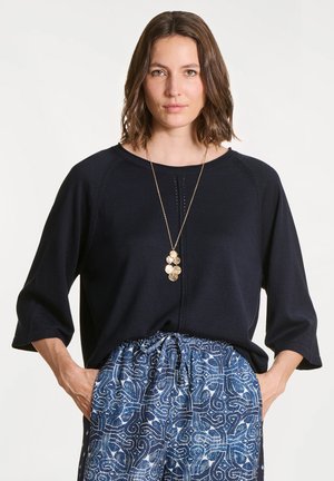 Femme portant un haut noir à manches trois quarts, un pantalon bleu à motifs avec cordon de serrage, et un collier pendentif en or, debout les mains dans les poches.