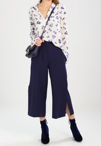 Blouse créme à fleurs avec des fleurs navy, accompagnée de culottes larges navy avec fentes sur les côtés. Le mannequin porte un petit sac bandoulière noir.