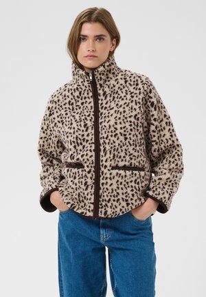 Junge Frau trägt eine Fleecejacke mit Reißverschluss in Beige und Dunkelbraun mit Leopardenmuster und blaue Jeans, steht mit den Händen in den Taschen.