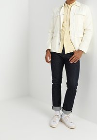 Leichte weiße Jacke mit mehreren Taschen, kombiniert mit einem blassgelben Hemd, dunkelblauen Jeans und weißen Sneakers, mit hochgerollten Ärmeln.