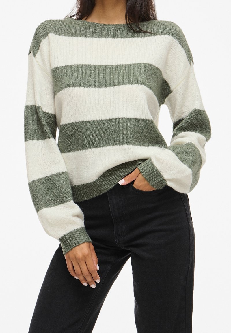 Femme portant un pull en maille à larges rayures vertes et blanches et un pantalon noir taille haute, une main dans la poche.