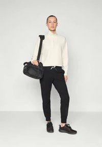 Hvit genser med glidelås i halsen, svarte joggebukser med snøring og svarte joggesko. Modellen holder en svart veske med skulderreim. Enkel, moderne design.