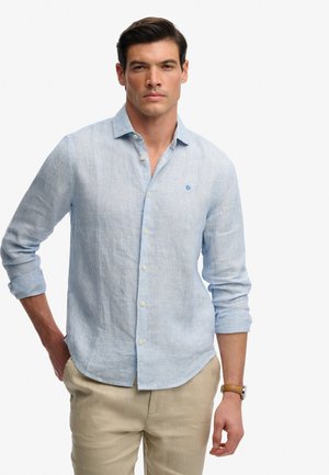 Mężczyzna ubrany w jasnoniebieską lnianą koszulę z guzikami i podwiniętymi rękawami oraz beżowe spodnie, stojący na jednolitym jasnym tle.