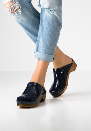 Granatowe, błyszczące klompy z drewnianymi podeszwami, zakrzywiony kształt i otwarty tył. Noszone z podwiniętymi jasnoniebieskimi, zniszczonymi dżinsami.