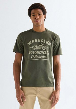 Junger Mann trägt ein olivgrünes Wrangler-T-Shirt mit Motorrad-Grafik und beigen Hosen, steht vor einem schlichten weißen Hintergrund.