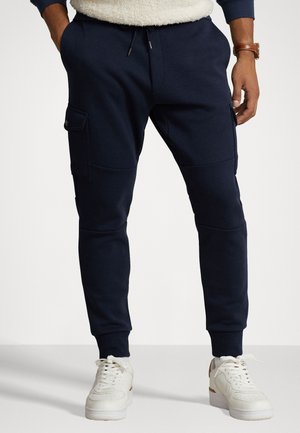 Mand iført marineblå joggingbukser med sidelommer, hvide sneakers og en brun ur på venstre håndled.
