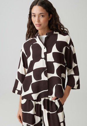 Frau trägt ein schwarz-weißes, abstrakt gemustertes Hemd mit Knöpfen und passende weite Hose, posiert mit einer Hand in der Tasche.