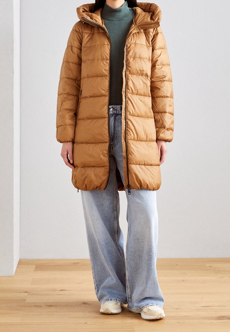 Veste matelassée marron avec capuche, fermeture éclair et motif matelassé, associée à un jean taille haute bleu clair et des baskets crème.