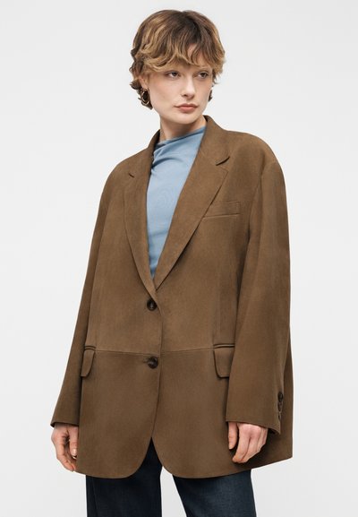 Personne portant un blazer marron surdimensionné par-dessus un haut bleu clair et un pantalon foncé, debout devant un fond uni gris clair.